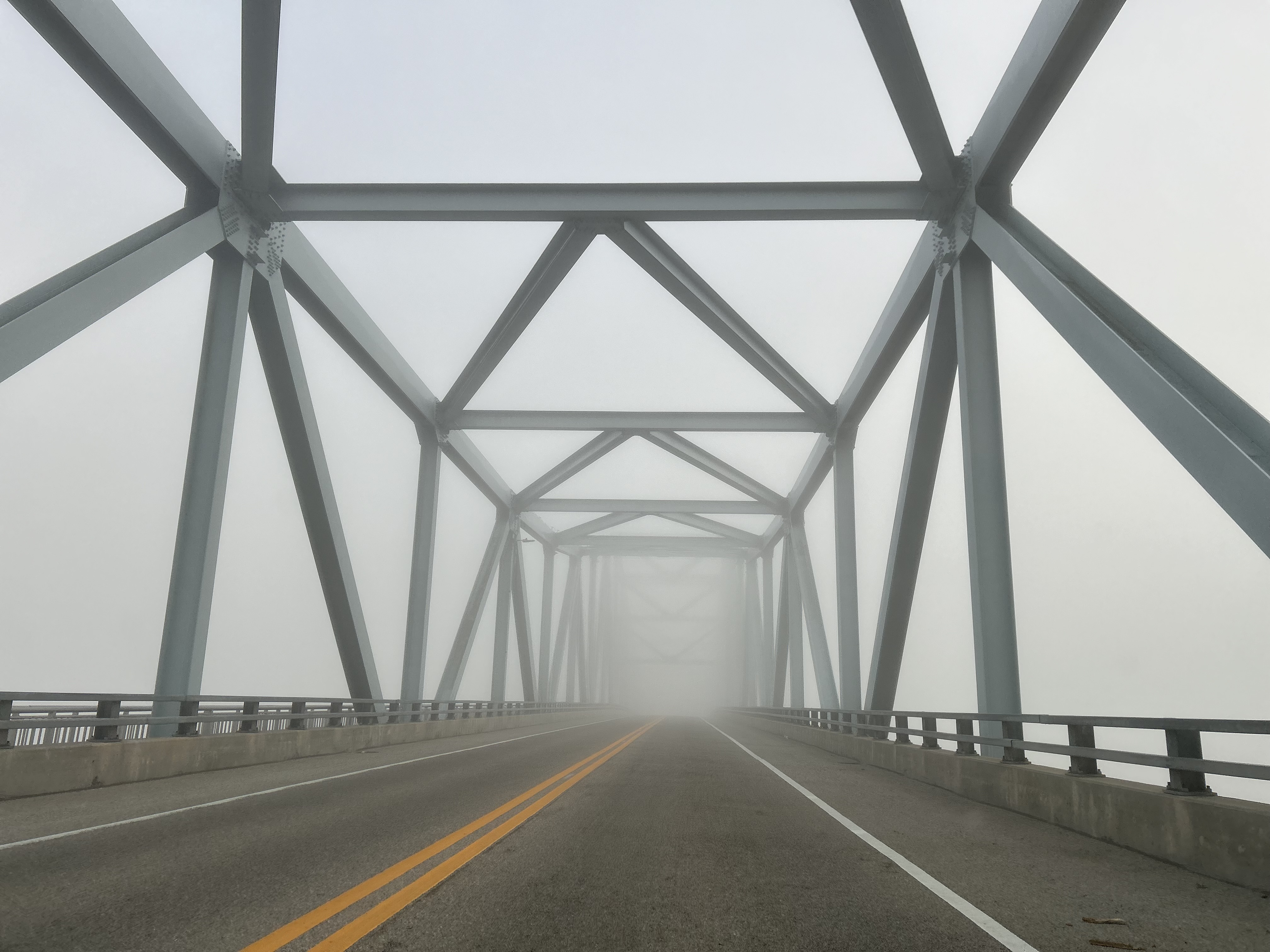 A long steel bridge with clean neutral gray tones stretches into perspective under overcast daylight. The symmetrical composition frames steel support beams leading toward a fog-shrouded vanishing point. Yellow center road lines and neutral gray railings guide the eye forward. The realistic, muted color palette and structural perspective convey a sense of clarity and direction.